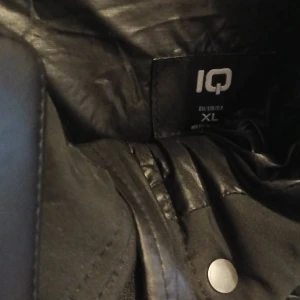 Svart pufferjacka från IQ - Säljer en svart pufferjacka från IQ i storlek XL. Jackan har en glansig yta med stora fickor och knappar framtill. Perfekt för kyliga dagar med sin vadderade design.