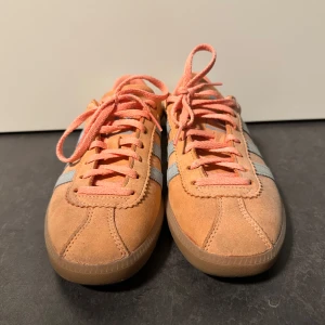 Adidas Gazelle i orange mocka - Snygga Adidas Gazelle sneakers i orange mocka med ljusblå ränder. Skorna har klassisk snörning och en bekväm gummisula. Perfekta för en avslappnad stil.