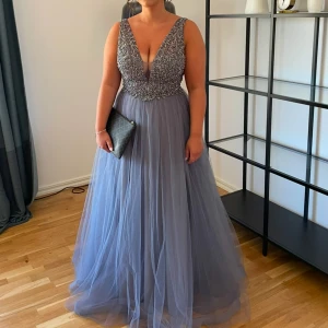 Blå långklänning med pärlor - Elegant blå långklänning med djup v-ringning och pärlbroderad överdel. Klänningen har en öppen rygg och är ärmlös, vilket ger en sofistikerad look. Perfekt för speciella tillfällen.