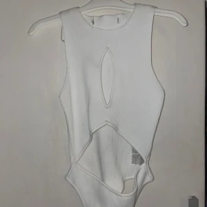 Vit ribbad ärmlös bodysuit - Snygg vit ribbad bodysuit med unika utskärningar framtill. Den är ärmlös och har en stilren design som passar perfekt för en trendig look. Perfekt att matcha med jeans eller kjol för en chic outfit.