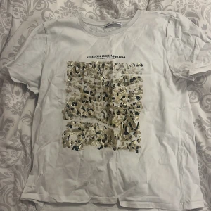 Vit t-shirt med leopardmönster från Zara - Säljer en vit t-shirt från Zara med ett coolt leopardmönster i guld och svart på framsidan. T-shirten är kortärmad och perfekt för en avslappnad stil. Inga defekter bara lite skrynklig vilket enkelt kan fixas. Passar bra till jeans eller shorts för en trendig look.