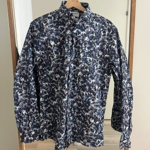 Blommig skjorta från Lindbergh - Snygg blommig skjorta från Lindbergh i blå och vit färg. Skjortan har långa ärmar och ett stilrent mönster som passar perfekt för en trendig look. Den är tillverkad i ett bekvämt material och har knappar framtill. Relaxed fit och superlex. Knappt använd 