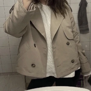 Beige kappa med knappar - Snygg beige kappa med dubbelknäppning och bred krage. Perfekt för en stilren look. Jackan har långa ärmar och en klassisk design som passar till många tillfällen.