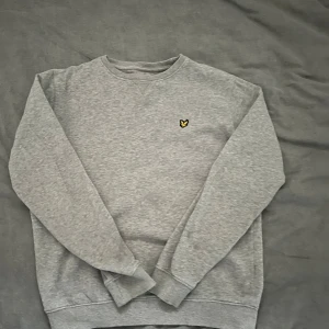 Grå sweatshirt från Lyle & Scott - Säljer en stilren grå sweatshirt från Lyle & Scott med deras ikoniska gula logga på bröstet. Tröjan har långa ärmar och en klassisk rund halsringning. Perfekt för en avslappnad stil.