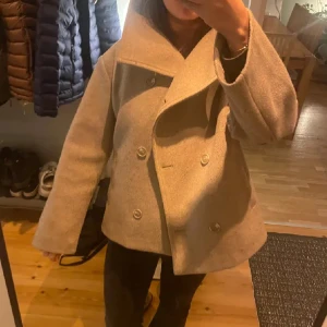 Beige kappa med dubbelknäppning - Snygg beige kappa med dubbelknäppning och bred krage. Jackan har en stilren design med långa ärmar och en lös passform. Perfekt för höst och vår.