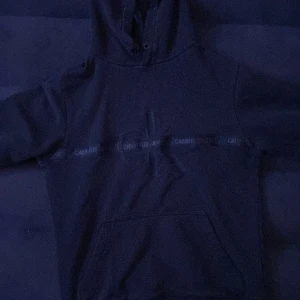 Svart hoodie från Calvin Klein Jeans - Säljer en stilren svart hoodie från Calvin Klein Jeans med deras ikoniska logga tryckt över bröstet. Hoodien har en klassisk design med känguruficka och justerbar huva. Perfekt för en avslappnad look.