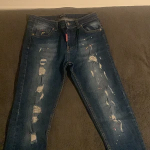 Dsquared2 jeans - Snygga blå jeans från Dsquared2 med coola slitningar och färgstänk. De har en klassisk femficksdesign och en röd logotypdetalj vid gylfen. Perfekta för en trendig look.