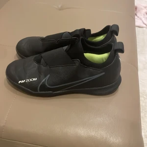 Svarta Nike fotbollsskor - Snygga svarta Nike fotbollsskor med Air Zoom-teknologi. Skorna har en stilren design med elastisk rem över vristen för extra stöd. Insidan är grön och yttersulan har bra grepp för inomhusspel.