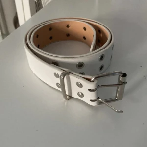 Vitt bälte med silverdetaljer - Snyggt vitt bälte med silverfärgad spänne och nitar. Perfekt för att ge en stilren touch till din outfit. Bältet har flera hål för justerbar passform.