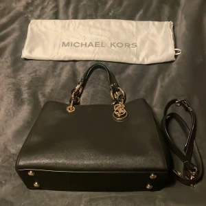 Svart handväska från Michael Kors - Snygg svart handväska från Michael Kors med gulddetaljer och en praktisk dragkedja. Väskan har ett elegant och stilrent utseende med ett avtagbart axelband. Perfekt för att bära dina nödvändigheter med stil.