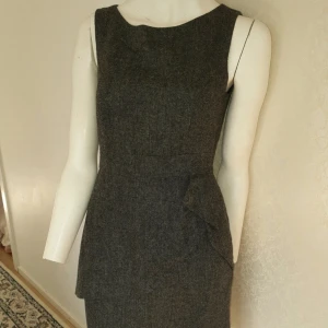 Grå ärmlös klänning - Snygg grå ärmlös klänning med en elegant volangdetalj på sidan. Klänningen har en diskret dragkedja i ryggen och är perfekt för en stilren look. Den är knälång och har en klassisk passform.