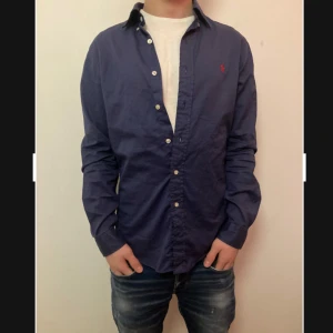 Mörkblå skjorta från Ralph Lauren - Snygg mörkblå skjorta från Ralph Lauren i slim fit. Skjortan har långa ärmar och knappar framtill. Perfekt för en stilren look med en liten röd broderad logga på bröstet. Feather weight twill-material för en lätt och bekväm känsla.