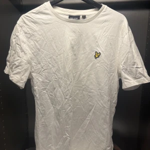 Vit t-shirt från Lyle & Scott - Säljer en klassisk vit t-shirt från Lyle & Scott med en liten broderad logga i gult och svart på bröstet. T-shirten är kortärmad och tillverkad i bomull, perfekt för en avslappnad stil.