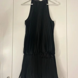 Svart plisserad klänning - Elegant svart plisserad klänning med knytning i nacken och dekorativa rosdetaljer i midjan. Perfekt för en stilren look.