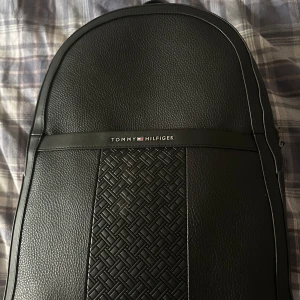 Svart ryggsäck från Tommy Hilfiger - Snygg svart ryggsäck från Tommy Hilfiger med ett stilrent flätat mönster på framsidan. Material läder. Väskan har justerbara axelremmar och en praktisk dragkedja. nypris är 1500. Säljer den för jag inte har använt den. Perfekt för att bära dina dagliga nödvändigheter med stil.
