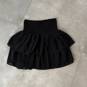 Svart volangkjol - Snygg svart kortkjol med volanger och en elastisk midja för bekväm passform. Perfekt för att skapa en stilren look med en touch av elegans. Kjolen har en luftig design som ger en fin rörelse.