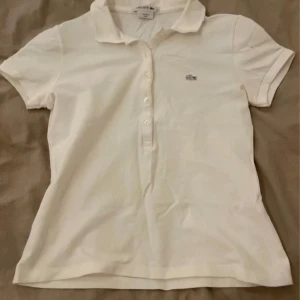 Vit kortärmad polotopp från Lacoste - Säljer en klassisk vit polotopp från Lacoste med korta ärmar och knappar framtill. Den har den ikoniska krokodilloggan på bröstet. Perfekt för en stilren och sportig look.