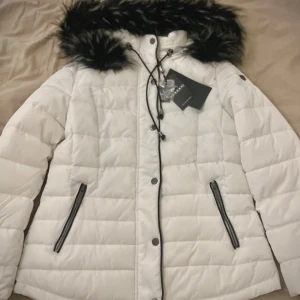 Vit dunjacka från Koröshi - Snygg vit dunjacka från Koröshi med svart fuskpäls på huvan. Jackan har dragkedja och knappar framtill samt två praktiska fickor med dragkedjor. Perfekt för kyliga dagar.