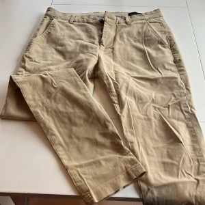 Beige chinos från Tailored Originals - 2 par Snygga beige chinos från Tailored Originals med klassisk design. De har en midwaist passform och stängs med dragkedja och knapp. Perfekta för en stilren look.