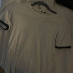 Vit t-shirt med svarta stickade detaljer - Säljer en stilren vit t-shirt från Zara med svarta kanter på ärmarna. Perfekt för en enkel och snygg look. T-shirten är kortärmad och gjord i ett mjukt material.