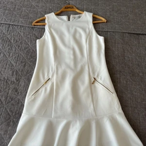 Vit klänning från Oxygene - Elegant vit klänning från Oxygene med en stilren design. Klänningen är ärmlös och har en dragkedja baktill. Den har dekorativa dragkedjor framtill och en volangdetalj vid fållen. Perfekt för en chic look!