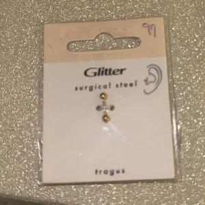 Tragus piercing i kirurgiskt stål från Glitter - Elegant tragus piercing i kirurgiskt stål från Glitter. Designad med en enkel stav och två guldfärgade kulor. Perfekt för att ge en subtil men stilren touch till din look.