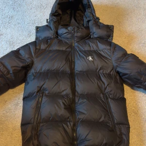 Svart dunjacka från Calvin Klein  - Säljer en snygg svart dunjacka från Calvin Klein med dragkedja och huva. Jackan har en avtagbar luva med dragkedja 