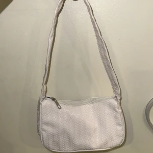 Vit axelväska med dragkedja - Snygg vit axelväska med ett diskret mönster och praktisk dragkedja upptill. Perfekt för att bära dina nödvändigheter med stil. Väskan har en bekväm axelrem som gör den lätt att bära med sig.