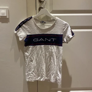 Vit t-shirt gant - Säljer en vit t-shirt  Gant med blått och rött band över bröstet och logotyp på ärmarna. 