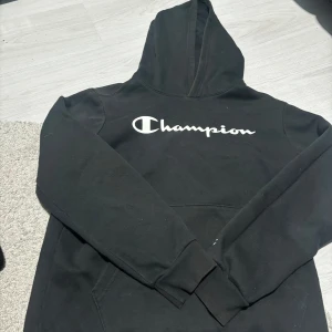 Svart hoodie från Champion - Säljer en klassisk svart hoodie från Champion med deras ikoniska logga tryckt på bröstet. Perfekt för en avslappnad stil med en stor ficka framtill och bekväm huva. Passar både för chill och aktivitet.