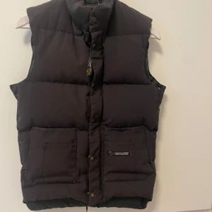 Svart dunväst från Canada Goose - Säljer en svart dunväst från Canada Goose har en hög krage och stängs med dragkedja och knappar. Den har två stora fickor framtill och en diskret logga på ena fickan. Perfekt för kyligare dagar.