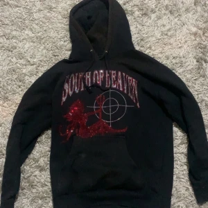 Svart hoodie från South of Heaven - Säljer en svart hoodie från South of Heaven med ett coolt rött glittertryck av en siluett och texten 'South of Heaven' på framsidan. Hoodien har en klassisk passform med långa ärmar och en praktisk ficka framtill. Perfekt för en avslappnad stil.