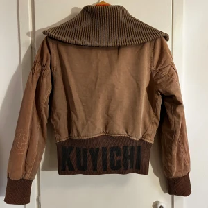 Brun jacka från Kuyichi - Säljer en snygg brun jacka från Kuyichi med stor ribbad krage och muddar. Jackan har dragkedja framtill och två fickor med knappar. Den har en cool textdetalj på ryggen och är perfekt för höst och vår då den är lätt fodrad🌟