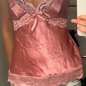 Rosa satinlinne med spetsdetaljer - Säljer ett vackert rosa linne i satin med spetsdetaljer vid halsringning och nederkant. Linnet har smala axelband och en liten rosett framtill. Perfekt för en elegant look.