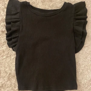 Svart ribbad volangtopp från Zara - Supersöt trendig svart ribbad volangtopp från Zara. Den är i storlek S men skulle definitivt säga att det mer är som en XS då den är lite tajtare. Jätte fint skick!❣️