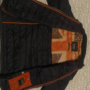 Mörk blå quiltad jacka från Barbour - Snygg svart quiltad jacka från Superdry med dragkedja och flera fickor med knappar. Insidan har ett coolt mönster med Union Jack. Perfekt för höst och vår!