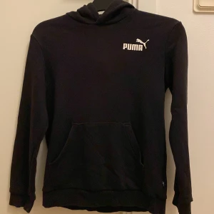 Svart hoodie från Puma - Säljer en svart hoodie från Puma med vit logga på bröstet. Tröjan har en klassisk design med huva och långa ärmar. Perfekt för en avslappnad stil.