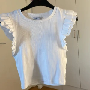Vit ribbad topp från Zara - Säljer en söt vit ribbad topp från Zara med volangdetaljer på ärmarna. Perfekt för en stilren och chic look. Toppen är ärmlös och har en rund halsringning.