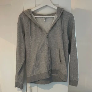 Grå hoodie med dragkedja - Säljer en klassisk grå hoodie med dragkedja och huva. Tröjan har långa ärmar och praktiska fickor framtill. Perfekt för en avslappnad stil. Hör av dig vid intresse!