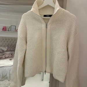 Vit stickad kofta från Bershka - Säljer en stilren vit stickad kofta från Bershka med dragkedja framtill. Koftan har en hög krage och långa ärmar, perfekt för kyligare dagar. Den är både mysig och trendig, ett måste i garderoben! 🤍