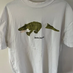 Vit t-shirt med krokodiltryck från Palm Angels - Säljer en vit t-shirt från Palm Angels med ett coolt krokodiltryck på framsidan och Palm Angels-logga på baksidan. Perfekt för en avslappnad stil. Nypris 2500