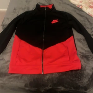 Svart och röd jacka från Nike - Säljer en snygg svart och röd jacka från Nike med dragkedja framtill. Jackan har en sportig look med långärmade ärmar och en hög krage. Perfekt för en casual stil eller träning.