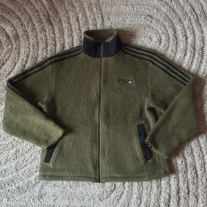 Adidas originals - Olivgrön fleecejacka (M)  - Säljer en sällsynt olivgrön/khaki fleecejacka (från Adidas originals =  SPRT Sherpa) med svarta detaljer och dragkedja. Jackan har de klassiska tre ränderna längs ärmarna och en broderad logotyp på bröstet, Perfekt för kyliga dagar!  Använt Max 10ggr.  Nästan Slutsåld överallt. Storlek: M [känns lite större och passar perfekt för L]  • Bröst: 93-100 cm Midja: 81-88 cm Höft: 92-99 cm • Material: 100% polyester. 