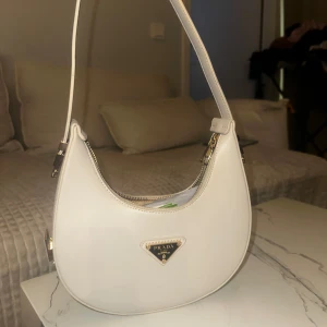 Vit axelväska från Prada - Snygg vit axelväska från Prada med en stilren design. Väskan har en dragkedja upptill och en justerbar axelrem. Perfekt för att ge din outfit en lyxig touch.