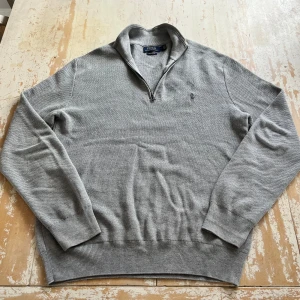 Grå tröja från Polo Ralph Lauren - Säljer en stilren grå tröja från Polo Ralph Lauren i mjuk Pima bomull. Tröjan har en halv dragkedja och ett diskret broderat märke på bröstet. Perfekt för en avslappnad men ändå snygg look.