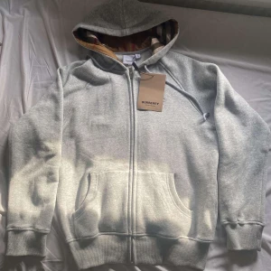 Grå hoodie från Burberry - Säljer en stilren grå hoodie från Burberry med dragkedja och broderad logga på bröstet. Den har en klassisk huva med snörning och ribbade muddar vid ärmslut och nederkant. Perfekt för en avslappnad look.