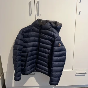 Mörkblå pufferjacka från Colmar - Säljer en snygg mörkblå pufferjacka från Colmar. Jackan har en praktisk huva och dragkedja framtill. Perfekt för kyliga dagar med sin quiltade design som håller värmen. Den har en stilren look med en diskret logga på ärmen.