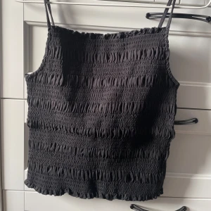 Svart smockad linnetopp - Snygg svart linnetopp med smockad design och tunna axelband. Passar bra till både jeans och kjol. Lappen är borta så jag vet inte vad det är för storlek men det ser ut som en S/M