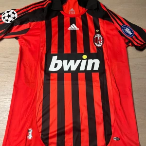 Röd och svart randig AC Milan fotbollströja - Säljer en klassisk AC Milan fotbollströja från Adidas med röd och svart randigt mönster. Tröjan har korta ärmar och Champions League-märke på ärmen. Den har även spelarnamnet 'Kaka' och nummer 22 på ryggen. Perfekt för samlare eller fans!