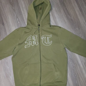 Olivgrön hoodie med tryck från tuttolente - Säljer en olivgrön hoodie med dragkedja och huva. Framsidan har ett tryck med bokstäverna 'MT' i vitt. Baksidan pryds av ett subtilt örntryck i samma färg som tröjan. Perfekt för en avslappnad stil. Xl men passar för L